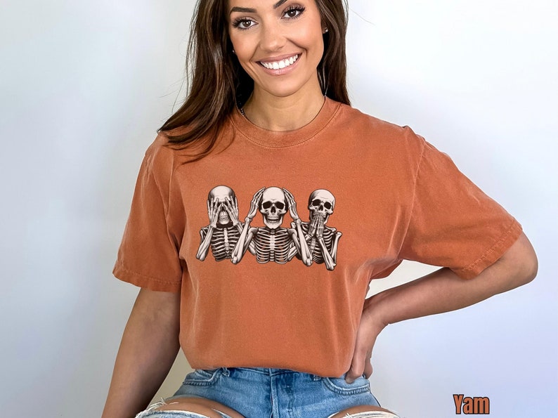 K&ouml;nnte beinhalten: Gebrannt orangefarbenes T-Shirt mit einer Grafik von drei Skeletten in der Pose &bdquo;nichts sehen, nichts h&ouml;ren, nichts sagen&ldquo;. Das Shirt hat eine lockere Passform und kurze &Auml;rmel. Das Wort &bdquo;Yam&ldquo; ist unten rechts aufgedruckt.