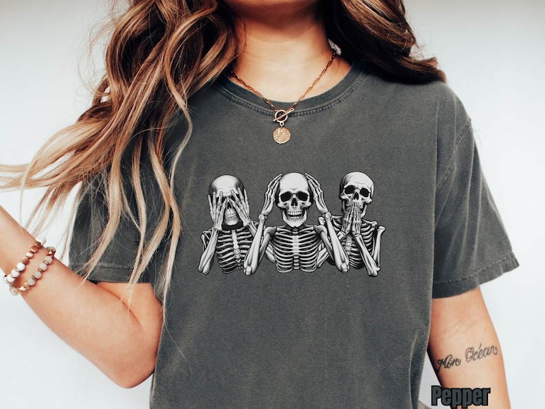 K&ouml;nnte beinhalten: Pfefferfarbenes T-Shirt mit einer Grafik von drei Skeletten in den Posen 'nichts sehen, nichts h&ouml;ren, nichts sagen'. Die Skelette sind in Schwarz und Wei&szlig; gehalten und weisen detaillierte Brustk&ouml;rbe und Sch&auml;del auf. Das Shirt hat eine lockere Passform.