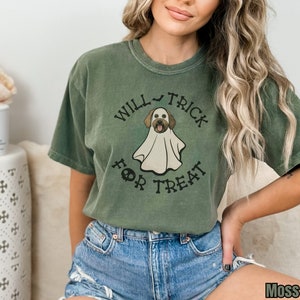 Puede incluir: Una camiseta de manga corta color verde musgo con el gr&aacute;fico de un perro vestido con un disfraz de fantasma blanco. El texto "WILL TRICK FOR TREAT" est&aacute; impreso en negro alrededor de la ilustraci&oacute;n, con un icono de murci&eacute;lago y una calavera.