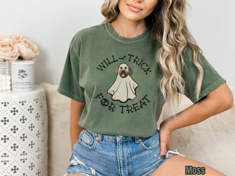 Puede incluir: Una camiseta de manga corta color verde musgo con el gr&aacute;fico de un perro vestido con un disfraz de fantasma blanco. El texto "WILL TRICK FOR TREAT" est&aacute; impreso en negro alrededor de la ilustraci&oacute;n, con un icono de murci&eacute;lago y una calavera.