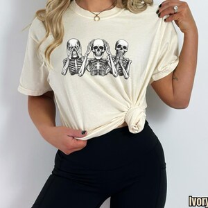 K&ouml;nnte beinhalten: Elfenbeinfarbenes T-Shirt mit einer schwarz-wei&szlig;en Grafik von drei Skeletten in der Pose &bdquo;nichts sehen, nichts h&ouml;ren, nichts sagen&ldquo;. Das Shirt hat kurze &Auml;rmel und einen Rundhalsausschnitt.