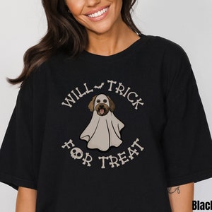 Puede incluir: Una camiseta negra con un gr&aacute;fico de un perro vestido con un disfraz de fantasma blanco. El texto "WILL TRICK FOR TREAT" est&aacute; impreso en una fuente con forma de hueso alrededor del perro, con un icono de murci&eacute;lago y una calavera.