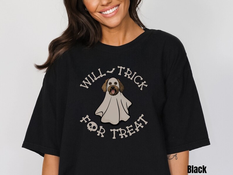 Puede incluir: Una camiseta negra con un gr&aacute;fico de un perro vestido con un disfraz de fantasma blanco. El texto "WILL TRICK FOR TREAT" est&aacute; impreso en una fuente con forma de hueso alrededor del perro, con un icono de murci&eacute;lago y una calavera.