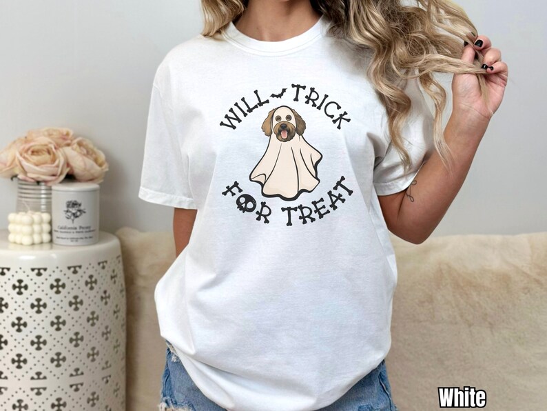 Puede incluir: Una camiseta blanca de manga corta con un gr&aacute;fico de un perro marr&oacute;n disfrazado de fantasma. El texto "WILL TRICK FOR TREAT" est&aacute; impreso en negro alrededor del perro, con un peque&ntilde;o icono de murci&eacute;lago arriba y una calavera debajo.