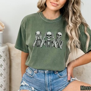 K&ouml;nnte beinhalten: Moosgr&uuml;nes T-Shirt mit kurzen &Auml;rmeln und Rundhalsausschnitt, das eine Grafik von drei Skeletten in der Pose &bdquo;nichts sehen, nichts h&ouml;ren, nichts sagen&ldquo; zeigt. Das Wort &bdquo;Moss&ldquo; ist unten rechts aufgedruckt.