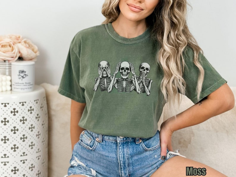 K&ouml;nnte beinhalten: Moosgr&uuml;nes T-Shirt mit kurzen &Auml;rmeln und Rundhalsausschnitt, das eine Grafik von drei Skeletten in der Pose &bdquo;nichts sehen, nichts h&ouml;ren, nichts sagen&ldquo; zeigt. Das Wort &bdquo;Moss&ldquo; ist unten rechts aufgedruckt.