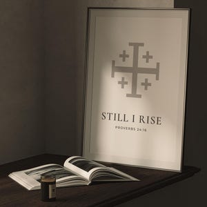 May include: A framed vertical wall art print featuring a gray Jerusalem cross symbol centered above the black text "STILL I RISE" and "PROVERBS 24:16" in a serif font. The print is displayed in a thin black frame with a light-colored background.