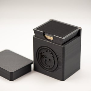 May include: A black, 3D-printed card box with a lid, and a separate lid. The box has a dragon emblem on the side. The lid is open, revealing the cards inside. The box is designed to hold playing cards.