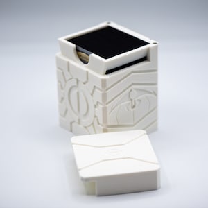 May include: A white, geometric box with a removable lid. The box is open, revealing a stack of black cards inside. The box has a detailed, textured design with geometric and stylized shapes. The lid is separate and rests in front of the box.
