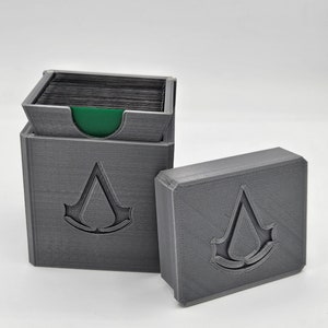 May include: Two grey 3D-printed card storage boxes. One box is open, revealing a stack of black cards and a green interior. The other box is closed. Both boxes feature the Assassin's Creed logo.