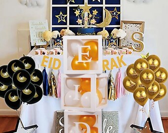 May include: A white box with clear sides filled with balloons that spell out "EID" in gold letters. The box is on a table with black and gold balloons, a gold banner that says "RAK", and a star-shaped light.