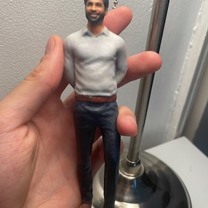 Könnte beinhalten: Eine 3D-gedruckte Figur eines Menschen, der ein weißes Hemd, einen braunen Gürtel und eine dunkelblaue Hose trägt. Die Figur hängt an einer silbernen Kette.