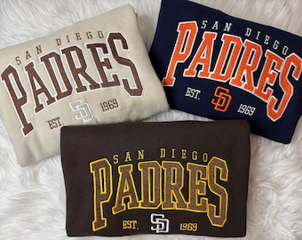 May include: Three folded sweatshirts featuring embroidered San Diego Padres text and logos. One is beige with brown text, one is navy blue with orange text, and one is dark brown with yellow text. Each includes the text EST. 1969 and a team logo.