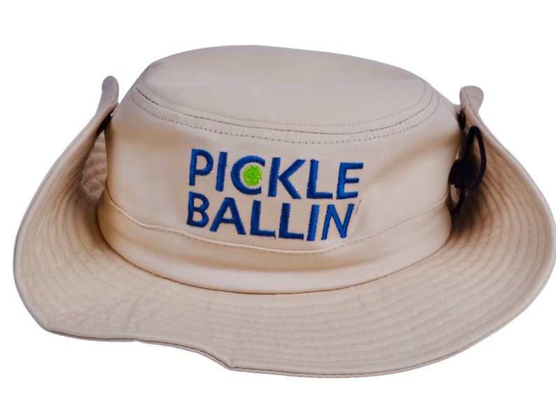 May include: A tan, wide-brimmed sun hat with a blue and green embroidered logo that says "PICKLE BALLIN".