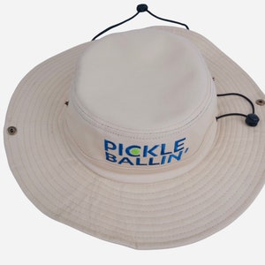 May include: A beige sun hat with a black drawstring and a green and blue embroidered logo that says "Pickleballin'"