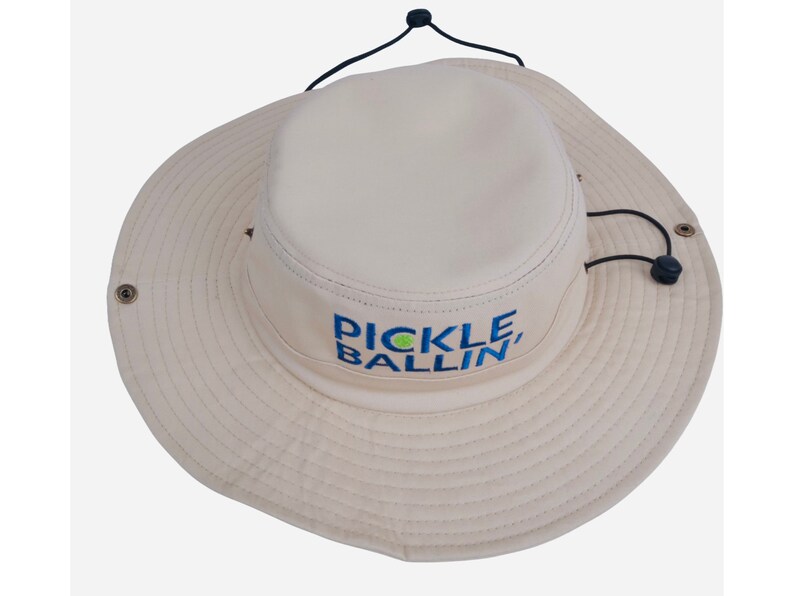May include: A beige sun hat with a black drawstring and a green and blue embroidered logo that says "Pickleballin'"