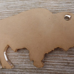 May include: A brown wooden bison cutout with a hole in the top for hanging. The cutout is on a light brown wooden surface.