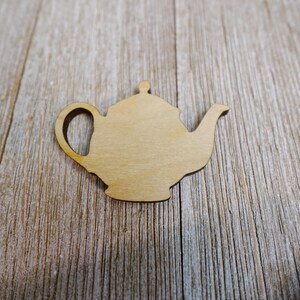 May include: A wooden teapot cutout with a handle, spout, and lid. The teapot is light brown and sits on a wooden surface. The cutout is a craft supply for DIY projects or decorations.
