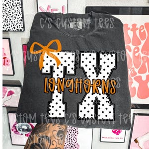 May include: A charcoal grey short-sleeve t-shirt featuring a large graphic of the letters TX in white with black polka dots. An orange bow sits atop the T, and the word Longhorns is printed in orange cursive script across the centre of the letters.