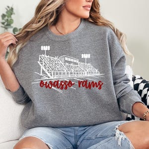May include: Grey sweatshirt with a white stadium graphic and the words "Owasso Rams" in red script. The sweatshirt has a crew neck and is made of a soft material. The design has a sporty theme.
