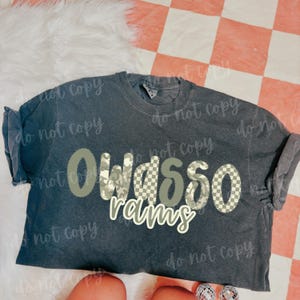 May include: Dark grey t-shirt featuring the text "Owasso Rams" in camouflage and checkered patterns. This casual wear item is likely for a sports team or school, suitable for everyday wear.