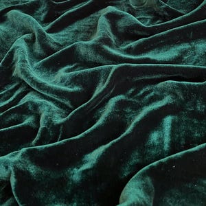 Puede incluir: Primer plano de una tela de terciopelo verde oscuro con una textura suave y lujosa. La tela está drapeada y doblada, creando un aspecto dramático y elegante.