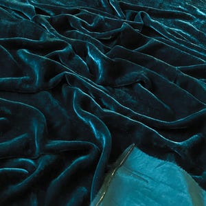 May include: Close-up of a teal blue velvet fabric with a soft, smooth texture. The fabric is draped and folded, creating a luxurious and elegant look.