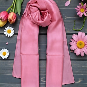 May include: A light pink silk scarf, draped on a dark gray wooden surface, with a soft, lustrous sheen. The scarf is surrounded by pink daisies, tulips, and white daisies, creating a floral accent. The scarf's rectangular shape is visible.