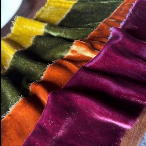 May include: Close-up of several strips of colorful velvet fabric. The colors include yellow, olive green, orange, and deep burgundy. The edges of the fabric are frayed, and the texture is soft and plush. Ideal for craft projects.