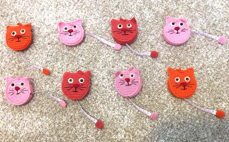 Puede incluir: Ocho cintas m&eacute;tricas de crochet con forma de gato. Cuatro son rosas y cuatro son naranjas. Cada cinta tiene una cara blanca, bigotes negros y ojos negros.