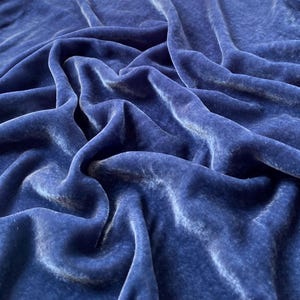 Può includere: Primo piano di un tessuto di velluto blu scuro con una texture morbida e vellutata. Il tessuto è drappeggiato e piegato, mostrando la sua sensazione di lusso.