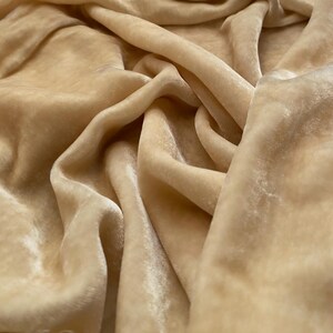 May include: A close-up of a soft, smooth, beige velvet fabric. The fabric is draped and folded, showcasing its texture and luxurious feel.