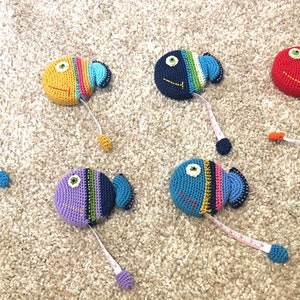 Puede incluir: Un conjunto de ocho cintas m&eacute;tricas de ganchillo con forma de pez en varios colores, incluyendo azul, rojo, naranja, amarillo y morado. Cada pez tiene un patr&oacute;n de color diferente y un ojo negro.