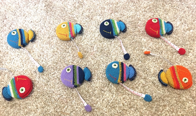 Puede incluir: Un conjunto de ocho cintas m&eacute;tricas de ganchillo con forma de pez en varios colores, incluyendo azul, rojo, naranja, amarillo y morado. Cada pez tiene un patr&oacute;n de color diferente y un ojo negro.
