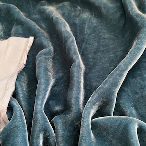 May include: Close-up of a teal blue velvet fabric with a soft, plush texture. The fabric is folded and draped, showcasing its luxurious feel.