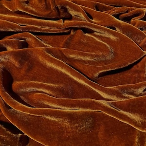 Puede incluir: Primer plano de una tela de terciopelo drapeada en un rico color marrón cálido. El material tiene una superficie suave y brillante, con pliegues y sombras que crean profundidad y textura. La tela parece gruesa y lujosa.