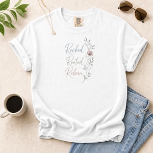 Può includere: Una t-shirt bianca a maniche corte con il testo Rocked Rooted Reborn in carattere corsivo, accompagnato da una delicata illustrazione botanica di foglie e un singolo fiore in tonalità tenui di grigio, blu e rosa tenue sul davanti.