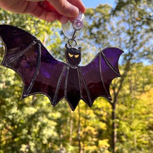 May include: A stained glass bat with purple wings and a black body. The bat has yellow eyes and is suspended by a suction cup. The bat is a Halloween decoration.
