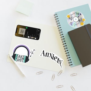 Puede incluir: Una imagen de plano de una superficie blanca con varios objetos, incluyendo un cuaderno con espiral, un cuaderno negro, una pantalla de teléfono que muestra una lista de reproducción de música, una pegatina de auriculares con el texto "Fake Shit", una pegatina con la palabra "Anxiety", y varios clips de papel.