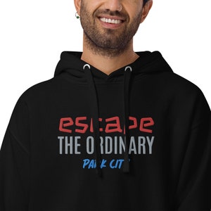 Op de afbeelding: Zwart hoodie met de tekst "escape the ordinary" in witte en rode letters en "park city" in blauwe letters.