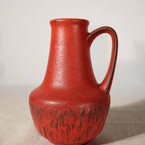 以下が含まれることがあります： テクスチャ加工された表面と茶色のハンドルが付いた赤いセラミックの花瓶。花瓶は丸みを帯びたボディと狭い首を持っています。