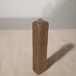 May include: A tall, rectangular brown ceramic vase with a slightly textured surface. The vase has a small, irregular opening at the top. The vase is displayed on a neutral-coloured linen surface.