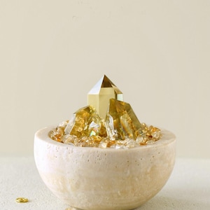 May include: A decorative arrangement featuring golden crystals in a beige stone bowl. The bowl is filled with small, amber-colored crystals, with larger, faceted crystals rising from the center. Small gold accents are scattered around the bowl.