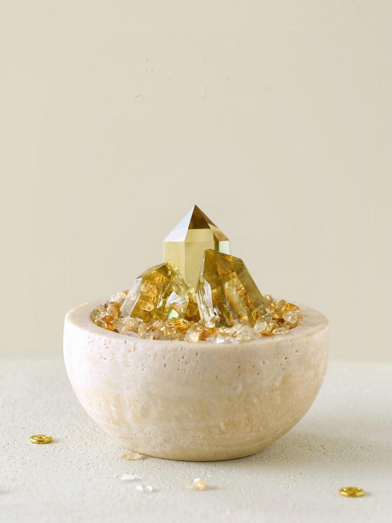 May include: A decorative arrangement featuring golden crystals in a beige stone bowl. The bowl is filled with small, amber-colored crystals, with larger, faceted crystals rising from the center. Small gold accents are scattered around the bowl.
