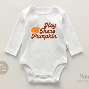 Peut inclure: Combinaison bébé à manches longues blanche avec une citrouille brune et le texte "Hey There Pumpkin" en brun.