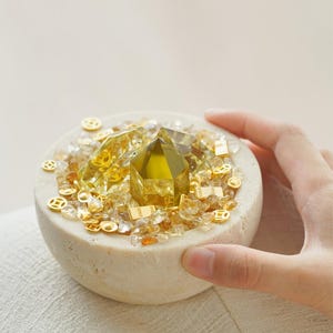 May include: A small, round, beige stone bowl filled with yellow and clear crystals, and small gold-colored decorative pieces. A large, faceted, yellow crystal is centered in the bowl. The bowl is held by a hand.