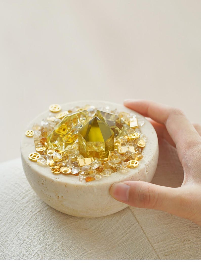 May include: A small, round, beige stone bowl filled with yellow and clear crystals, and small gold-colored decorative pieces. A large, faceted, yellow crystal is centered in the bowl. The bowl is held by a hand.