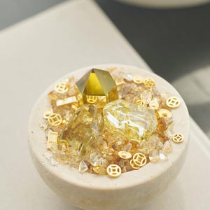 May include: A beige, round bowl filled with yellow and clear crystals, gold coins, and a large, faceted yellow crystal. The bowl has a rough texture and is set against a neutral background. The arrangement suggests a theme of wealth or prosperity.