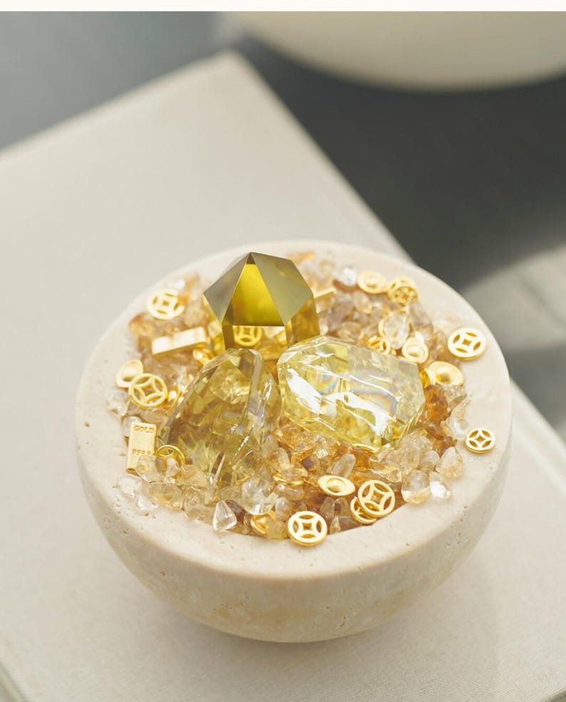 May include: A beige, round bowl filled with yellow and clear crystals, gold coins, and a large, faceted yellow crystal. The bowl has a rough texture and is set against a neutral background. The arrangement suggests a theme of wealth or prosperity.