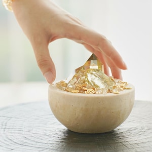 May include: A beige, semi-spherical bowl filled with golden crystals and small, decorative gold pieces. A large, faceted crystal sits in the center. A hand hovers over the bowl, and a beaded bracelet is visible in the background.